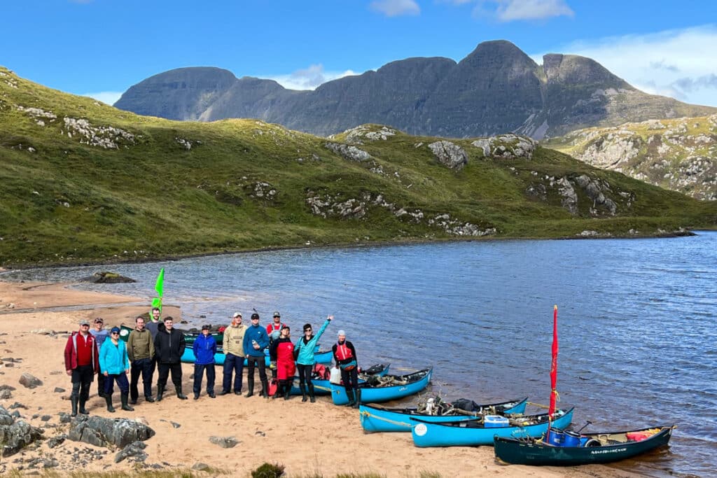 a picture of a group of people standing on a remote beach by their canoes, with wild mountainous scenery in background