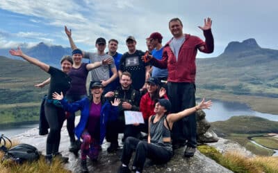 work colleagues on team building expedition wave from the summit of a hill, with wild mountainous landscape in the background.