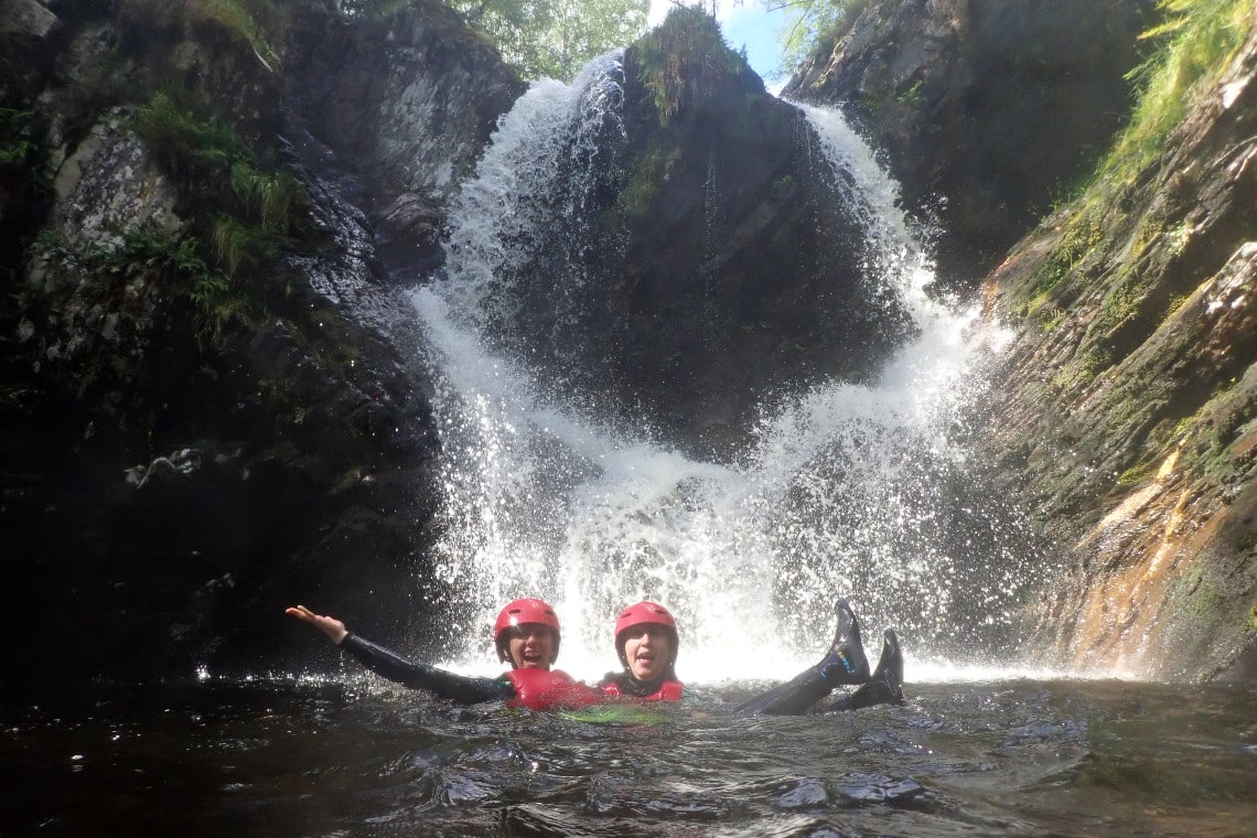 2 friends smiling and having a great time as they pose in front of a big waterfall. they are in the water in wetsuits and helmets, below the falls.