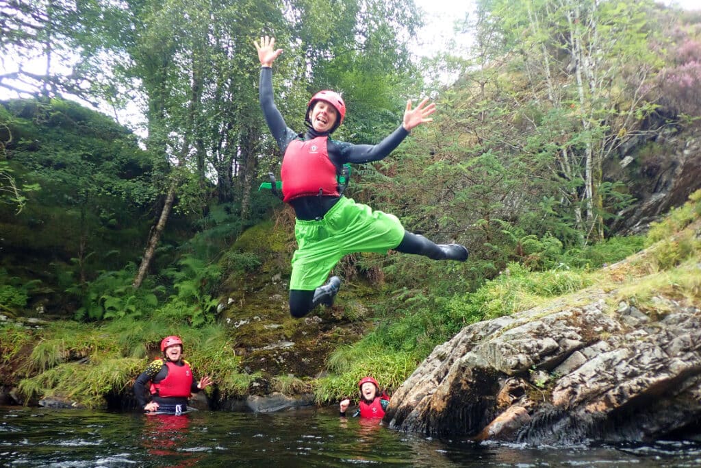 a female wiht big smile on her face making a big jump of a rock into the water while gorge walking in a highland glen.