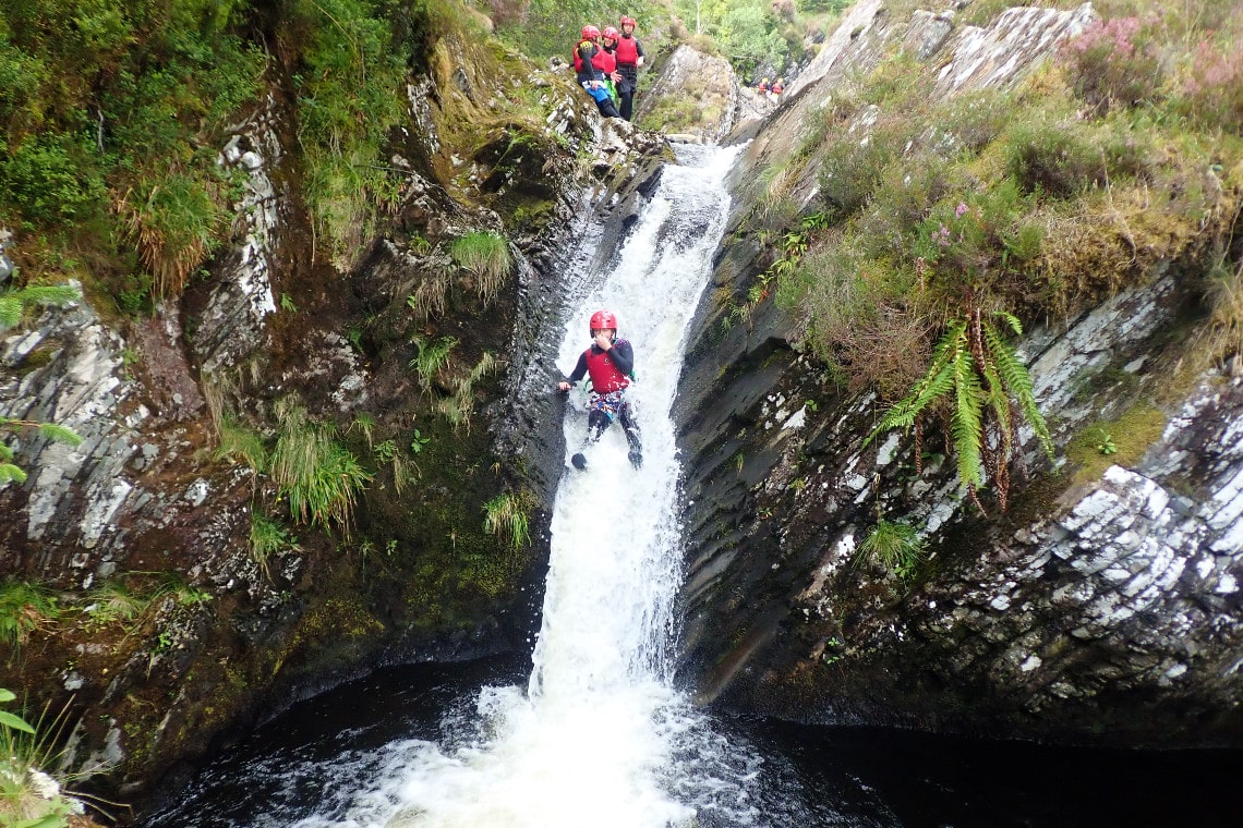 a person sliding down a big waterfall in wetsuit and helmet while gorge walking in a highland gorge