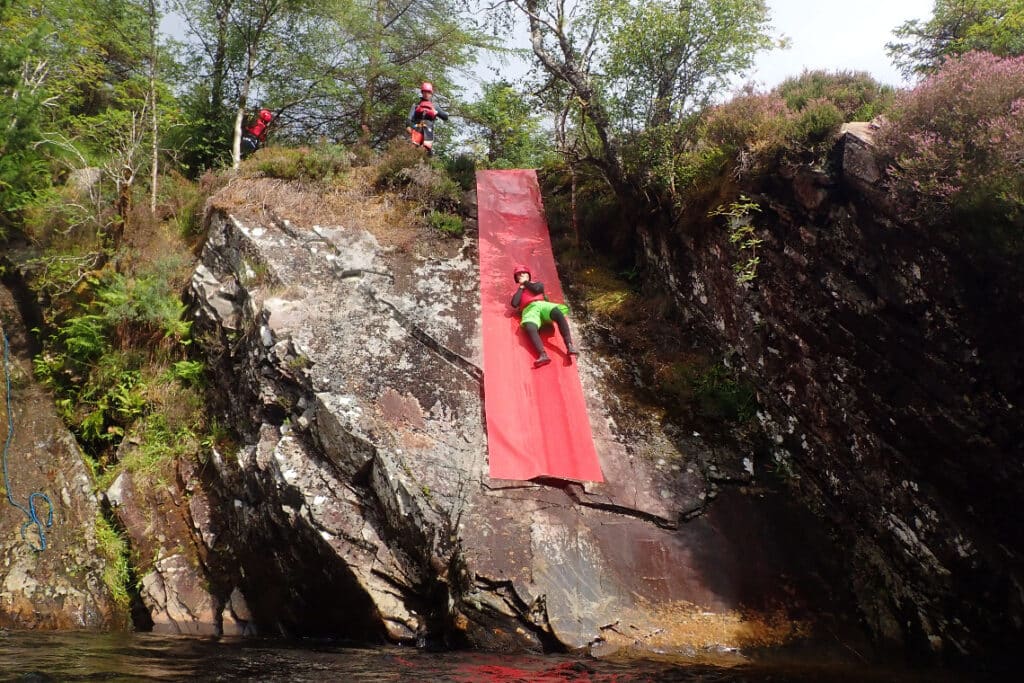 A person sliding on long red tarpaulin down a big rock face into a deep pool, while gorge walking in a scottish highland river gorge