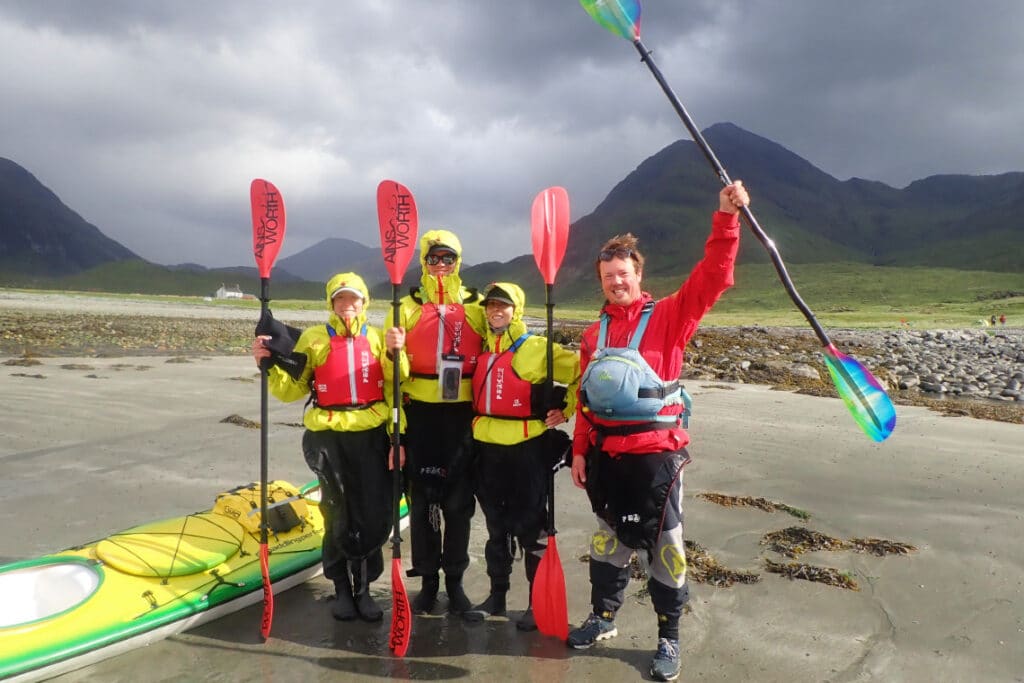 kayakers, holding paddles aloft, pose for a photo on the beach, next to their kayaks, waterproofs on, ready for the weather