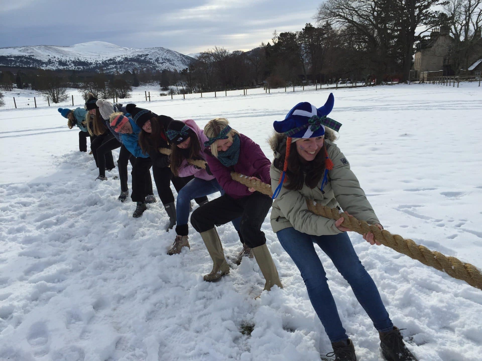 Group of women doing tug of war in the snow, with snowy hills in the background.